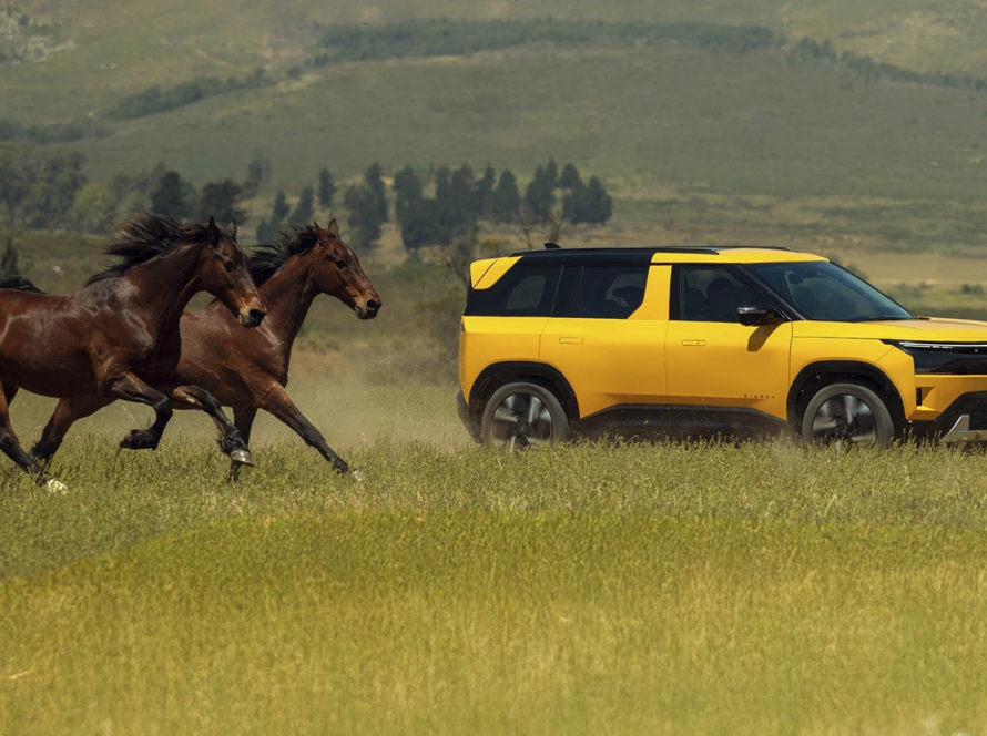 A yellow and black dual-tone new-generation Tata Sierra SUV driving on an outdoor, grassy plain with some horses galloping behind it.